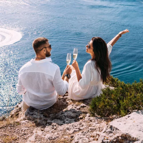 Couple sits on cliff above blue waters dressed in white with champagne flutes